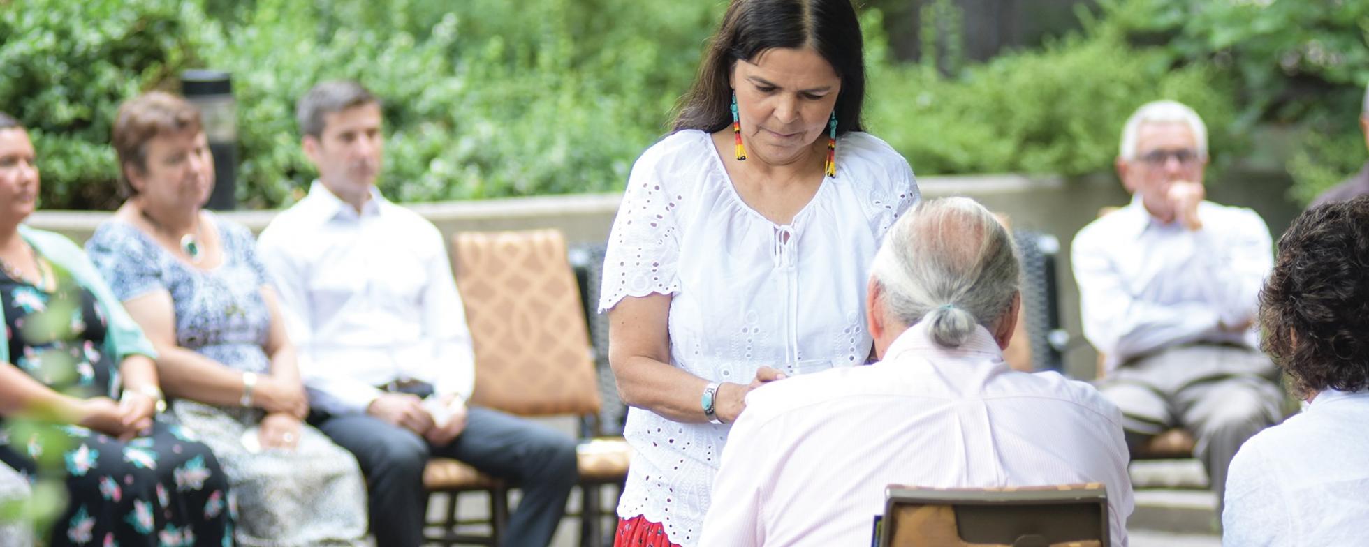 EN: A women is standing in front of a seated elderly man FR: Une femme se tient devant un homme âgé assis